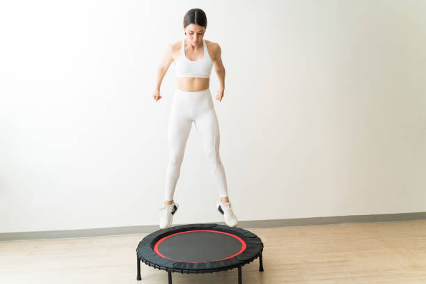 Pretty Young Woman In White Sportswear Jumping On Trampoline Attractive slim Caucasian female rebounding against wall during interval training at fitness studio trampoline stock pictures, royalty-free photos & images