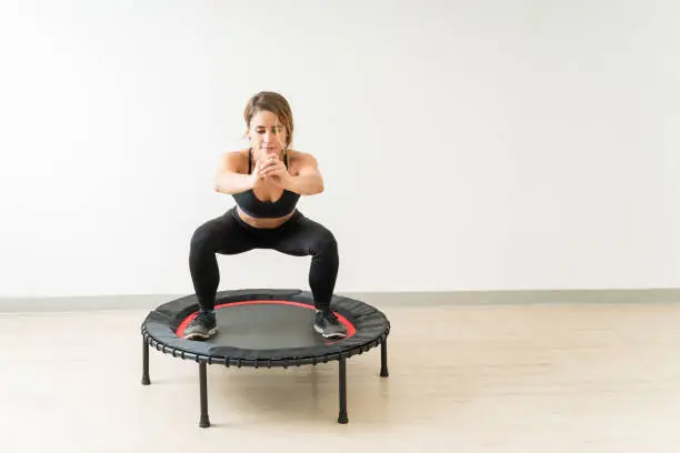 Pretty young brunette woman trampolining while doing squats against wall at studio during fitness training workout