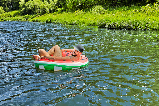 Young attractive caucasian man with hat on his head is drifting down the river in forest on the inflatable ring.