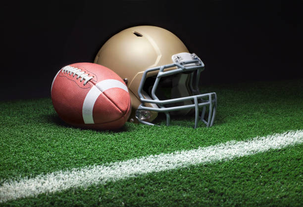 Low angle view of gold helmet and football on field with dark background A low angle view of a gold football helmet with a football on a grass field with stripe and dark background face guard sport photos stock pictures, royalty-free photos & images
