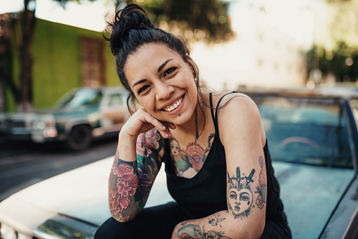 Tatuada Latina sonriendo al aire libre y sentada en la capucha del coche photo