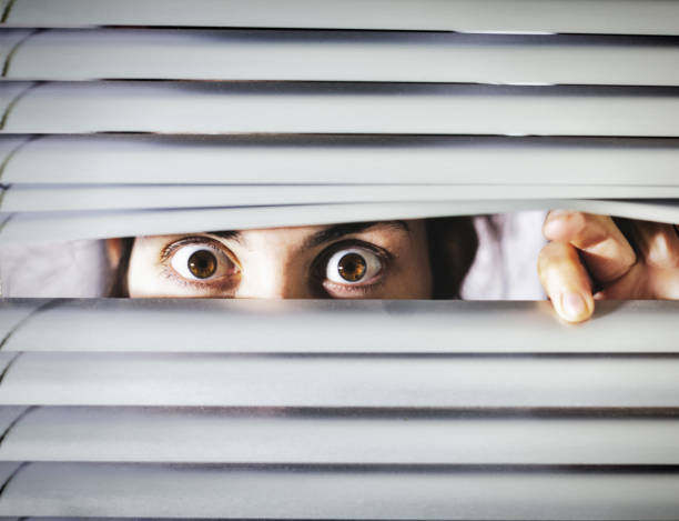 istock Wide-eyed young woman looking through blinds, terrified 1168130183