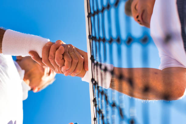 istock Men shaking hands while standing by tennis net against clear sky on sunny day 1168138004
