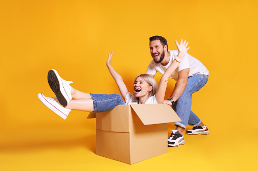 Young couple in matching outfits having fun moving boxes, isolated on yellow.