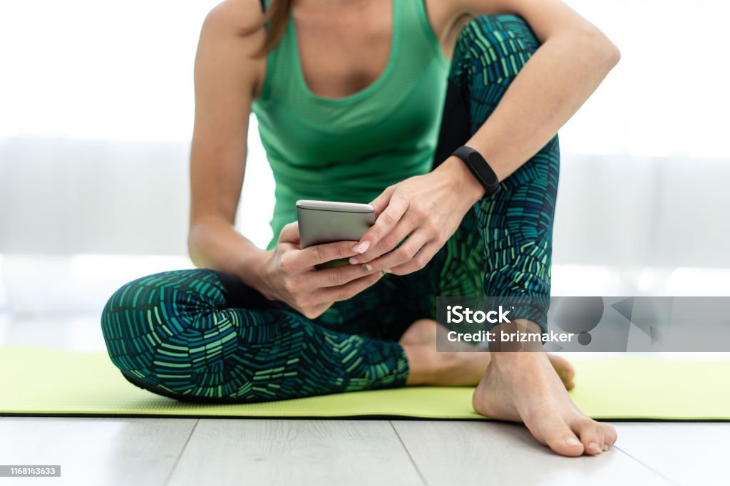 Young sporty woman using smartphone, sitting on fitness mat Cropped view of young woman sitting on fitness mat and using smartphone with mobile app after training at home Mobile App Stock Photo