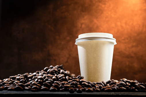 Styrofoam cup with hot coffee and coffee beans on the table over brown background