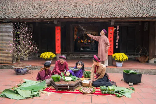 Vietnamese family members making Banh Chung together on old-styled house yard. Chung cake is a very well-known dish that could never miss on the altar, and family meal of Vietnamese during Tet holiday