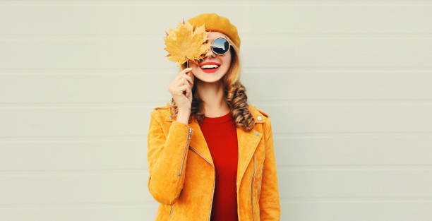 ¡estado de ánimo de otoño! mujer sonrisa feliz sosteniendo en sus manos hojas de arce amarillo escondiendo su ojo sobre el fondo gris de la pared - sunglasses women smiling portrait fotografías e imágenes de stock