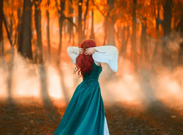 Photo of girl with bright red wavy hair sanding with back to camera, lady in green cloak