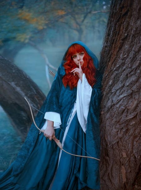 istock red-haired girl in blue cloak standing by tree and looking directly at camera 1168155510