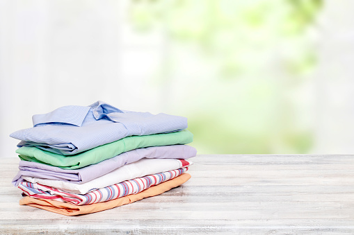 Stack colorful clothes. Pile of folded cotton shirts on a bright table with space for your display product montage against abstract blurred natural light green background. Summer fashion.