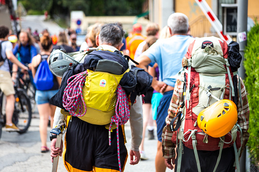 Chamonix, France - 5 August, 2019: color image depicting a rear view of two mountain climbers together walking on the street in the mountain resort of Chamonix in southern France close to the French Alps. The climbers are both carrying lots of gear, including heavy backpacks, climbing rope and helmets. They are dressed in full climbing clothing. Focus on the climbers with the street and tourists defocused in the background.