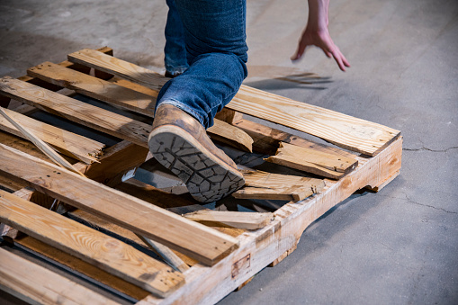 An industrial warehouse, workplace safety topic.  A close-up of a person stepping on a broken pallet that poses a severe risk of causing a fall and injury.