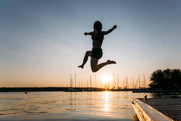 istock Summer day: young woman jump from jetty into lake while sunset 1168180583