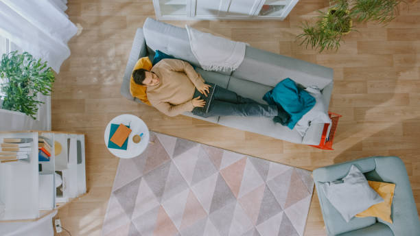 istock Young Man in Brown Jumper and Grey Jeans Lying Down on a Sofa, Using a Laptop. He is Happy and Smiles. Cozy Living Room with Modern Interior with Plants, Table and Wooden Floor. Top View. 1168183072