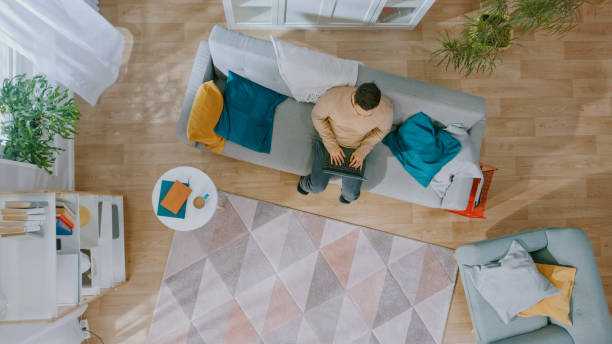 istock Young Man in Brown Jumper and Grey Jeans Comes and Sits Down on a Sofa, Using a Laptop. Cozy Living Room with Modern Interior with Plants, Table and Wooden Floor. Top View. 1168183077