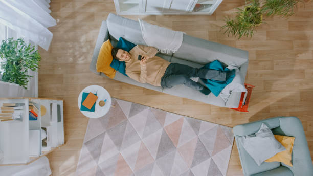 istock Young Man in Brown Jumper and Grey Jeans Comes and Lies Down on a Couch, Using a Smartphone. He is Happy and Smiles. Cozy Living Room with Modern Interior with Plants, Table and Wooden Floor. Top Down 1168183078