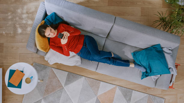 junges mädchen in rotem pullover und blue jeans liegt auf einem sofa, mit einem smartphone. sie ist glücklich und lächelt. gemütliches wohnzimmer mit modernem interieur mit teppich, couchtisch und holzboden. top-ansicht - carpet floor lying down people stock-fotos und bilder