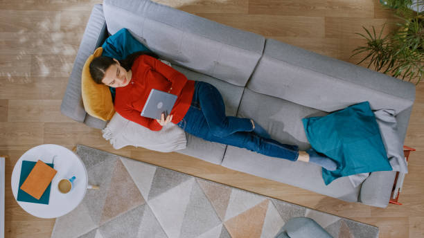 istock Young Girl in Red Blouse and Blue Jeans is Lying Down on a Couch, Using a Tablet Computer. She is Happy and Smiles. Cozy Living Room with Modern Interior with Carpet, Table and Wooden Floor. Top View. 1168183168