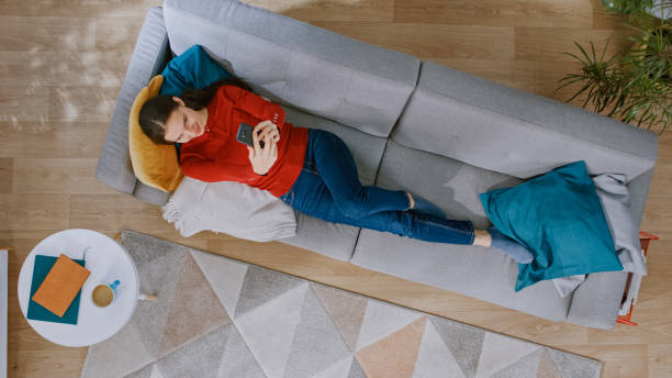 istock Young Girl in Red Jumper and Blue Jeans is Lying Down on a Sofa, Using a Smartphone. She is Happy and Smiles. Cozy Living Room with Modern Interior with Carpet, Coffee Table and Wooden Floor. Top View 1168183196