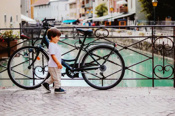 Photo of Little boy walking over bridge of Thiou River in Annecy, France near parked bicycle. Child in the city. Little tourist, summer lifestyle. Discovering Europe, traveling with kid.