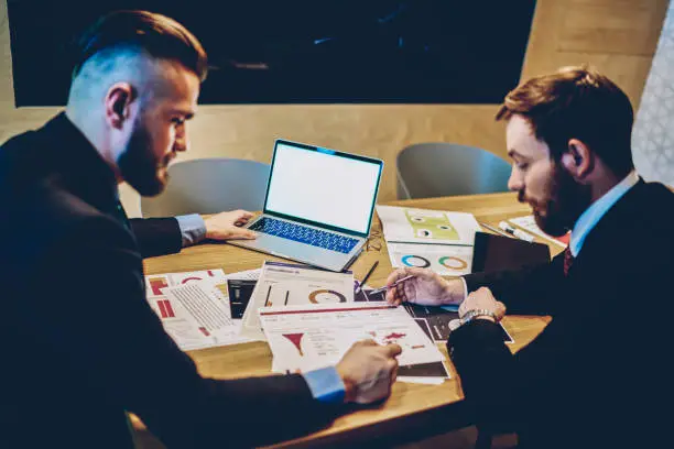 Male entrepreneurs at desktop with  graphics accountings discussing business plan for project, professional formally dressed economists checking results of campaign near laptop with blank screen