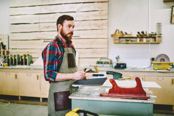 istock Pensive bearded handyman in apron standing with notepad pondering on organizing day in workshop, serious owner of manufacture business making notes about clients and orders in restoration store 1168188533