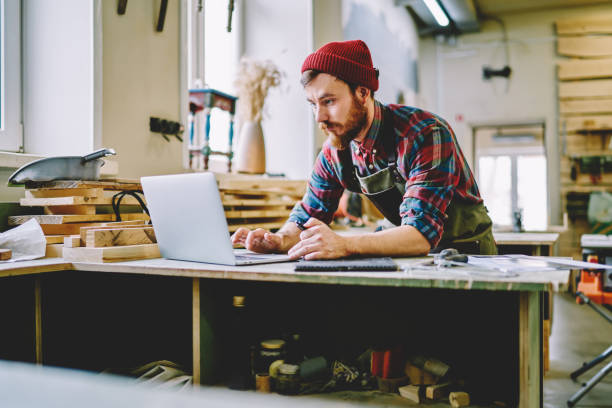 young hipster guy watching training tutorial on laptop computer learning at carpentry courses, skilled male artisan using netbook for searching information during woodworks and making plannings - training computer learning computer lab imagens e fotografias de stock