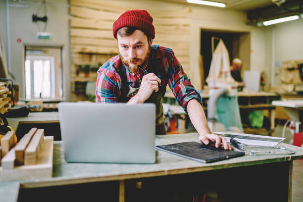 Serious bearded male master checking information from website while creating sketch and plannings for wooden stuff,skilled hipster guy watching training  webinar and making notes while learn on course