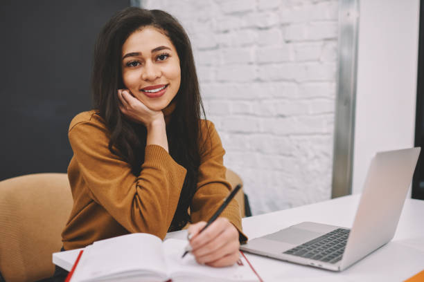 istock Portrait of smiling female student satisfied with learning at university making with modern technologies, skilled young woman writer looking at camera while making notes for her publication in office 1168197014