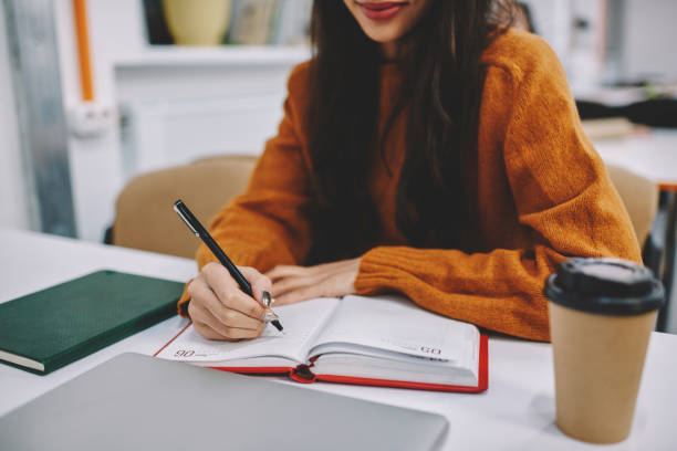 Cropped image of talented woman student making notations to notepad while preparing for coursework project sitting in campus, talented creative hipster girl recording information using black pen