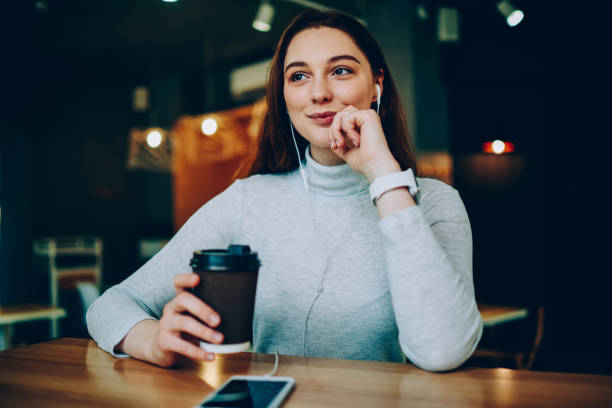 giovane donna sognante che si gode una nuova canzone negli auricolari durante la trasmissione radiofonica su smartphone, una ragazza hipster premurosa che beve caffè e ascolta un interessante audiolibri al telefono mentre riposa - the thinker audio foto e immagini stock