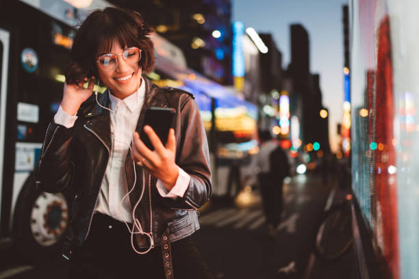 fröhliche hübsche junge frau in coolen brille und trendige tragen zu fuß auf metropole straße mit nachtlichter genießen audio-songs von playlist in kopfhörern mit smartphone-gerät verbunden - lab coat women one person female stock-fotos und bilder