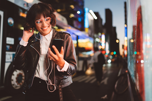 Cheerful pretty young woman in cool eyeglasses and trendy wear walking on metropolis street with night lights enjoying audio songs from playlist in earphones connected to smartphone device