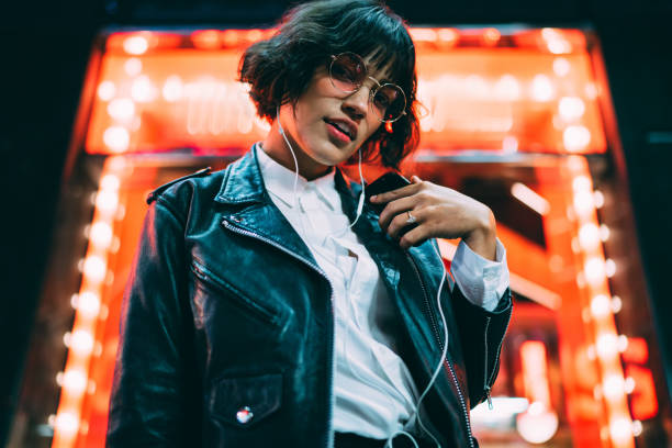 Half length portrait of attractive hipster girl dressed in fashionable apparel listening music in earphones from mobile player enjoying night lights in downtown.Stylish female meloman standing in neon illumination retrato de media longitud de atractiva chica hipster vestida con ropa de moda escuchando música en auriculares de un jugador móvil disfrutando de luces nocturnas en el centro de la ciudad. elegante meloman femenino de pie en iluminación de neón