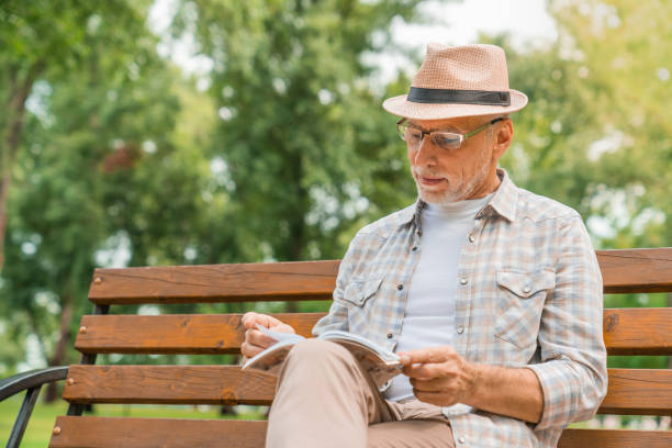 istock Senior man in hat reading book while sitting on public bench in park zone 1168207785