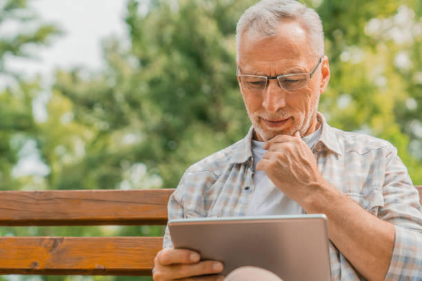 Handsome mature man in eyeglasses using digital tablet on bench in park Park, Digital Tablet, Mature Men, Beard, One Person reading newspaper stock pictures, royalty-free photos & images