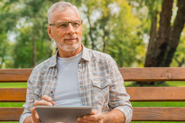 istock Handsome mature man in eyeglasses using digital tablet on bench in park with look away 1168208514