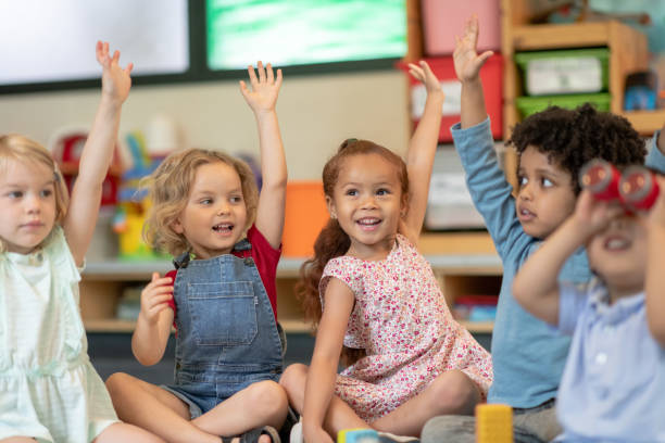 Multi-ethnic group of students in class A multi-ethnic group of preschool students is sitting with their legs crossed on the floor in their classroom. The teacher is sitting on the floor facing the children. The happy kids are smiling and listening to the teacher. They have their arms raised and are eager to answer the teacher's question. children only stock pictures, royalty-free photos & images