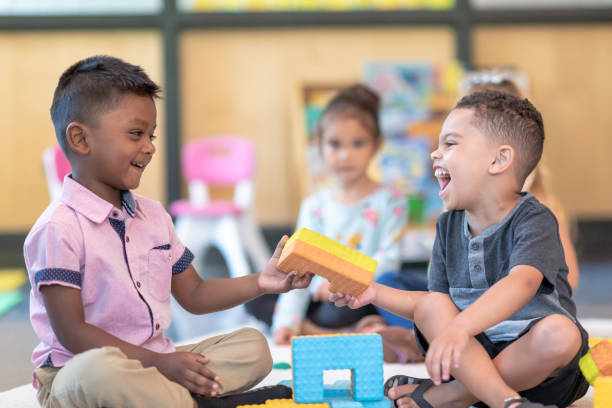 Happy preschool students playing together in classroom A multi-ethnic group of preschool students sits together on the floor in their classroom. The classmates are happily playing together. The focus is on two little boys who are laughing while building with toy blocks. Other children are playing in the background. montessori stock pictures, royalty-free photos & images