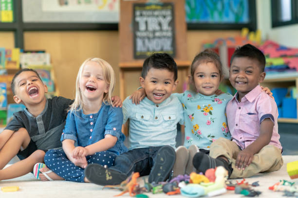 Group of smiling preschool students Portrait of a happy multi-ethnic group of preschool students in their classroom. The cute children are sitting in a line with their arms around each other. The kids are laughing and smiling directly at the camera. community center photos stock pictures, royalty-free photos & images