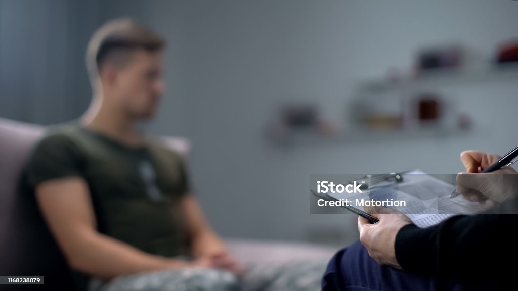 Psychologist making notes during therapy session with sad male soldier, PTSD Mental Health Professional Stock Photo