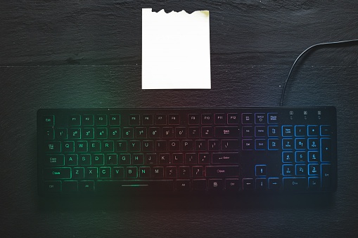Rgb computer keyboard with small piece of blank paper next to it with copy spade - Colorful modern device used for inserting data with notebook sheet on dark desk