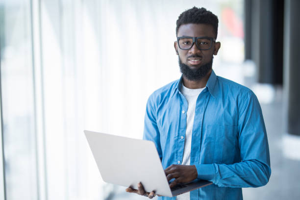 istock Excellent mood. Cheerful young african manager in glasses is working on laptop while sitting on table in modern office. He is looking at monitor with smile. 1168257714