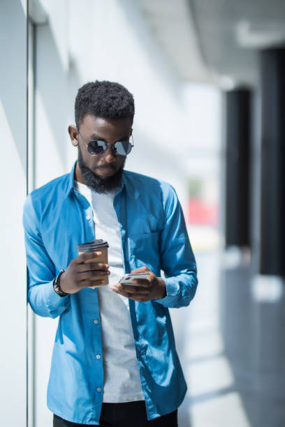 istock Young african-american businessman use mobile phone near panoramic windows in office while drink coffee 1168259898