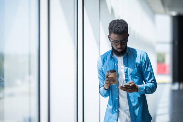 Young african-american businessman use mobile phone near panoramic windows in office while drink coffee