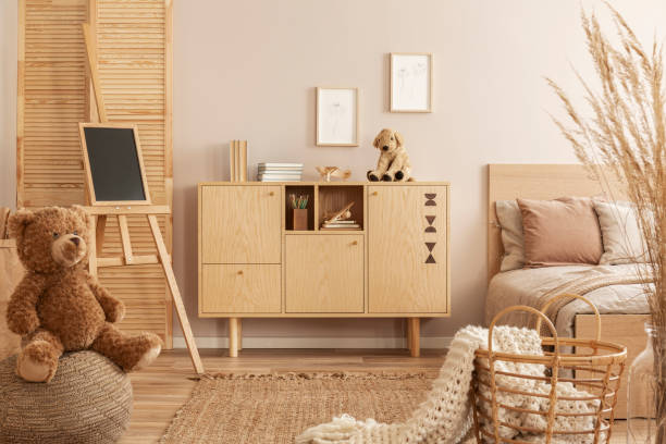 Teddy bear on pouf and small blackboard on easel in stylish kid's bedroom with wooden cabinet and beige bedding on bed Teddy bear on pouf and small blackboard on easel in stylish kid's bedroom with wooden cabinet and beige bedding on bed beige bedroom stock pictures, royalty-free photos & images