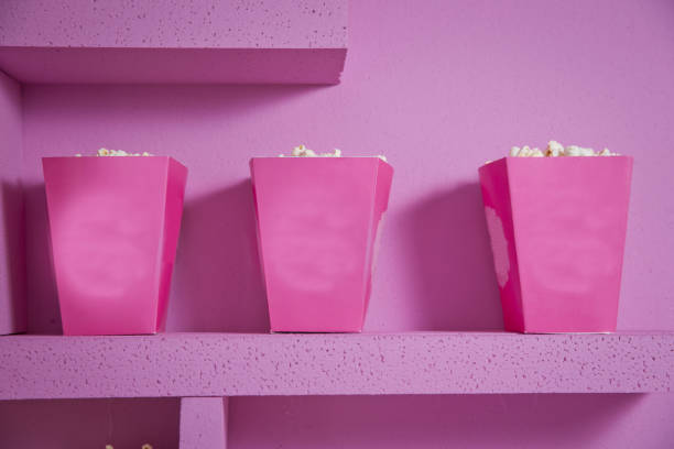 A wide classic box of theater popcorn isolated on pink . Popcorn spilled from a striped box . Three vintage-style boxes of popcorn in a carnival themed still life with arcade game pink tickets palomitas de maíz derramadas de una caja a rayas. tres cajas de estilo vintage de palomitas de maíz en un bodegones temático de carnaval con entradas rosas juego de arcade. una amplia caja clásica de palomitas de maíz de teatro aisladas en rosa.