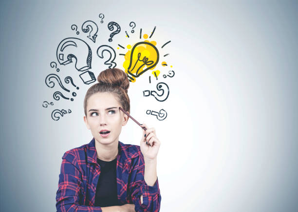 Thoughtful teen girl in casual clothes sitting near gray wall with question marks and lightbulb drawn on it. Concept of good idea. Mock up
