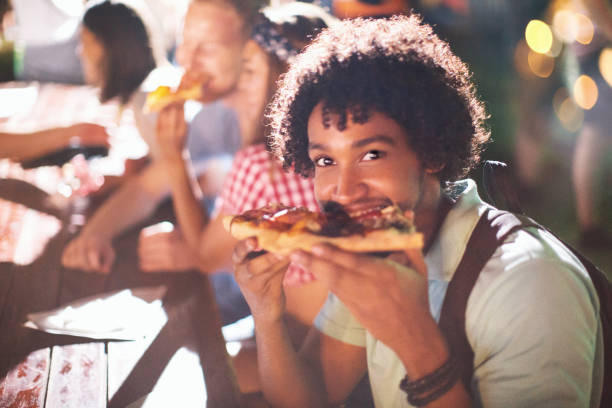 After party food time. Young african american man eating pizza after party. He is smiling and looking at camera. food festival stock pictures, royalty-free photos & images